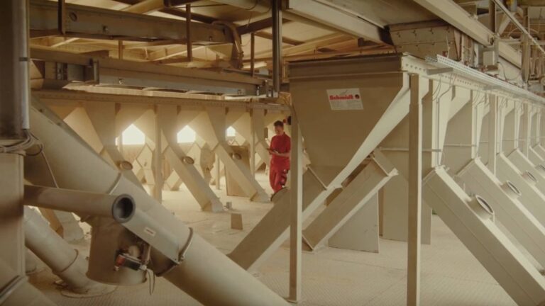 Mitarbeiter in roter Arbeitskleidung steht in einer industriellen Anlage mit Trichtern und Rohrsystemen zur Verarbeitung von Schüttgut.