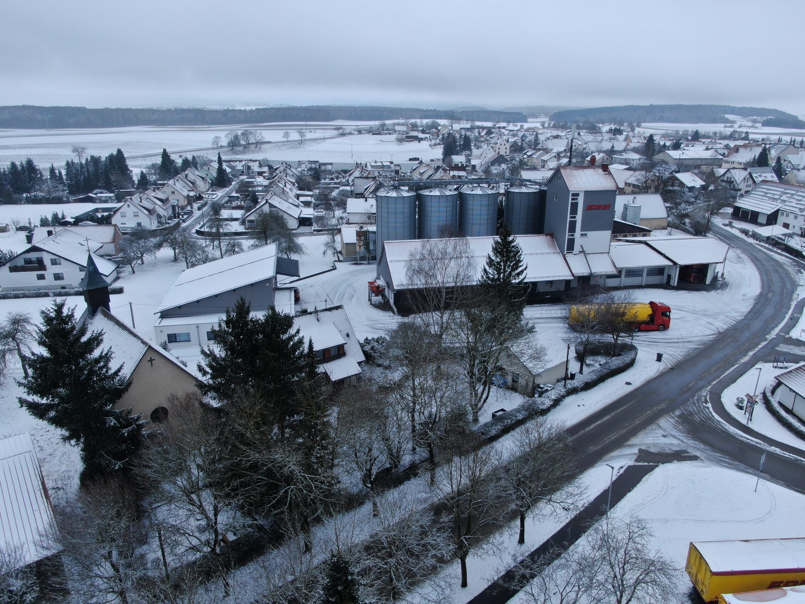 Luftaufnahme eines verschneiten Dorfes mit Agrarbetrieb, Silos und Lkw bei winterlicher Landschaft.